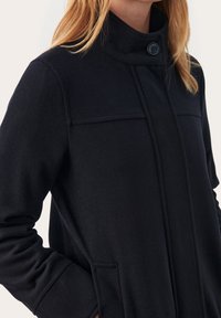 Sort uldfrakke med højt krave, enkelt knaplukning og sidelommer, præget af minimalistisk design og glat tekstur.