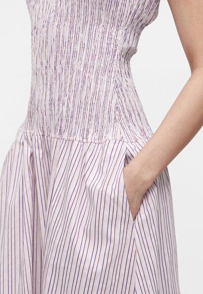 Χέρι γυναίκας στην τσέπη φορέματος χωρίς μανίκια, εφαρμοστό, με ραφή τύπου smock, με κάθετες μοβ ρίγες σε λευκό φόντο.