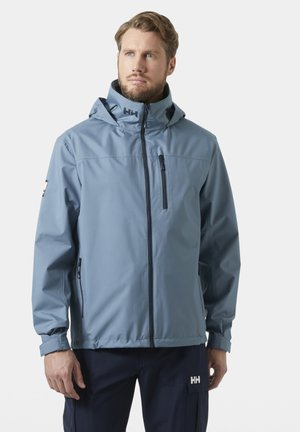 Hombre con chaqueta con capucha azul claro y pantalones oscuros, de pie y mirando ligeramente hacia un lado sobre un fondo liso.