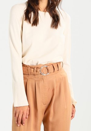 Vrouw die een crèmekleurige top met lange mouwen en geribbelde manchetten draagt, gecombineerd met een beige broek met hoge taille, een stoffen riem en een zilveren ronde gesp.