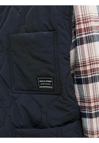 Marineblå quiltet vest med en firkantet lomme og logo-patch, parret med en creme- og bordeauxfarvet ternet stof på højre side.