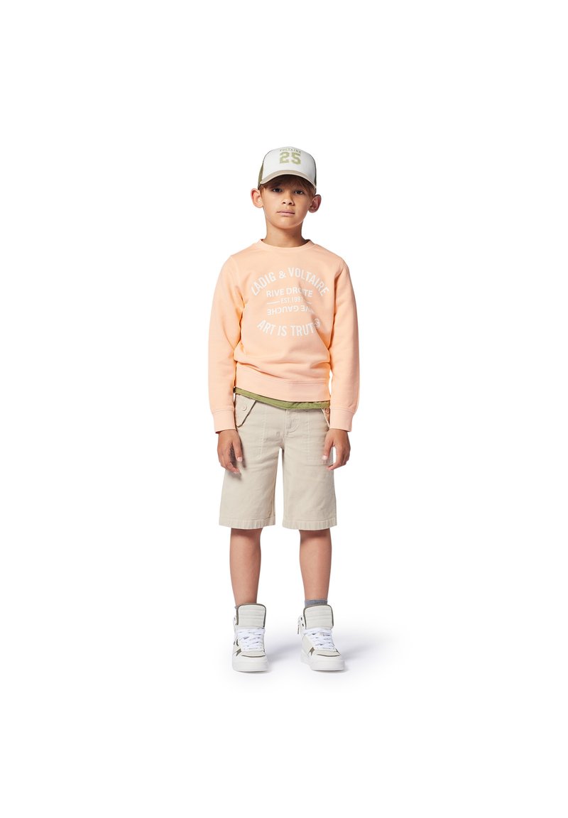 Lichtoranje sweatshirt met witte tekst, kaki shorts en witte hoge sneakers. Pet heeft een lichtgrijze kleur met groene accenten.