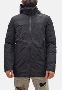 Giubbotto imbottito nero con zip frontale, cappuccio regolabile e tasche laterali. Presenta una texture liscia e opaca. Indossato sopra jeans strappati.