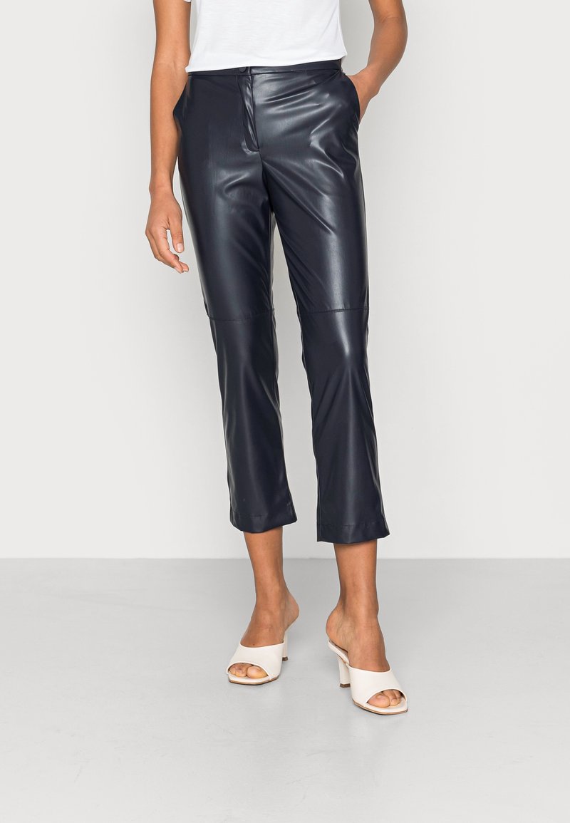 Pantalon court en cuir synthétique bleu marine avec une texture lisse et une silhouette ajustée. Associé à des talons ouverts crème.