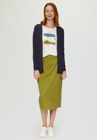 Jupe midi verte avec poches, assortie à un t-shirt graphique blanc et un cardigan marine. Le modèle porte des baskets blanches ; tissu lisse et coupe décontractée.
