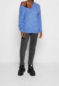 En person som bär en lös blå V-ringad tröja, kolgrå jeans och svarta kraftiga sneakers, står mot en helt vit bakgrund.