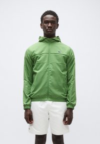 Zelena jakna z zadrgo in kapuco, s horizontalnimi šivi na prsih, elastičnimi manšetami in majhnim logotipom na levi strani prsnega koša; nošena z belimi kratkimi hlačami.