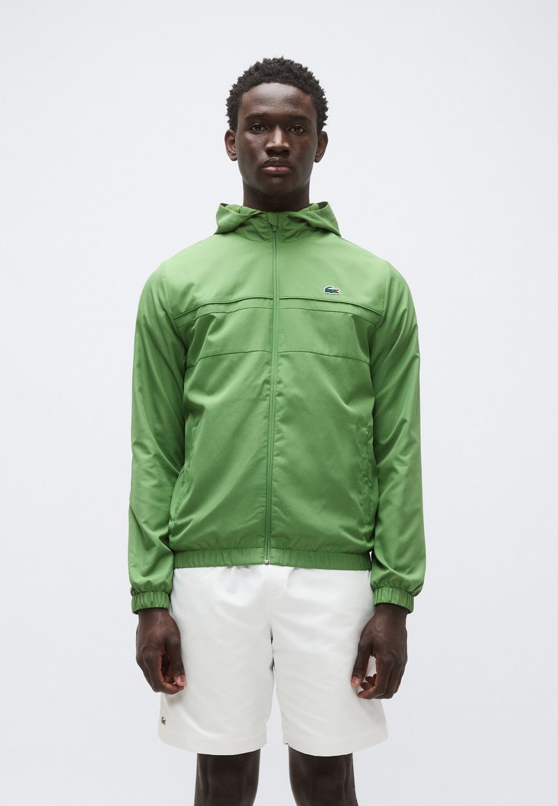 Zelena jakna z zadrgo in kapuco, s horizontalnimi šivi na prsih, elastičnimi manšetami in majhnim logotipom na levi strani prsnega koša; nošena z belimi kratkimi hlačami.