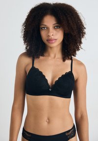 Svart blonde bralette med trekantet koppsdesign, bølgete kanter, justerbare stropper og en liten sølvdetalj i midten.