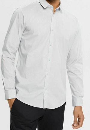 Hombre con una camisa blanca ajustada de botones y mangas largas y pantalones negros, mano en el bolsillo, contra un fondo liso.