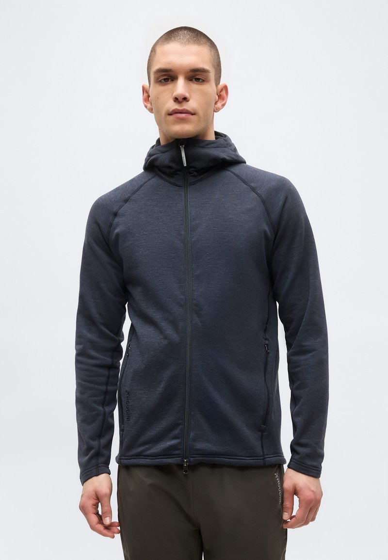 Νεαρός άνδρας στέκεται, φοράει μια σκούρα γκρι ζακέτα φερμουάρ με ψηλό γιακά και σκούρο παντελόνι σε απλό ανοιχτό φόντο.