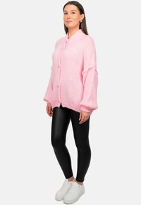 Roze gebreide cardigan met een sjawlboord en een sluiting met knopen aan de voorkant, gecombineerd met zwarte leggings en witte sneakers. Accenten omvatten losse mouwen.