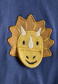 Sárga dinoszaurusz fejének hímzése, szarvakkal, kék textil háttérrel. Barna díszítésekkel és mosolygó arckifejezéssel.