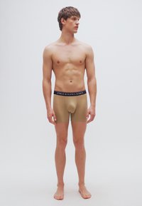 Μπεζ αντρικά εσώρουχα boxer με λουράκι μέσης σε ναυτικό μπλε χρώμα που φέρει το γράψιμο "POLO RALPH LAUREN". Η εφαρμογή είναι στενή με ένα λείο, ελαστικό ύφασμα.