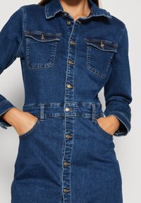 Jeansklänning i mörkblått med knappstängning framtill, krage, två bröstfickor och en figurnära midja. Har guldfärgade knapppdetaljer och långa ärmar.