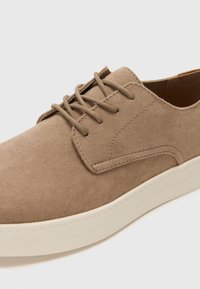 Beżowe zamszowe sneakersy o gładkiej fakturze, okrągłym czubku, minimalistycznym designie i kontrastowej kremowej gumowej podeszwie. Posiadają pasujące sznurowanie.