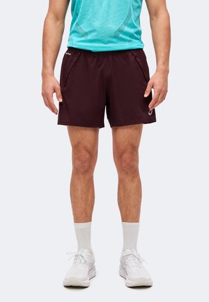 Personne portant un short de sport bordeaux foncé, des chaussettes blanches, des baskets blanches et un débardeur turquoise à motifs.