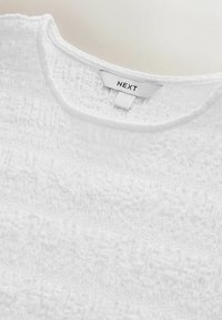 Haut blanc en tissu texturé, avec un col rond et l'étiquette de la marque "NEXT" à l'intérieur. Aucun accent ou motif notable.