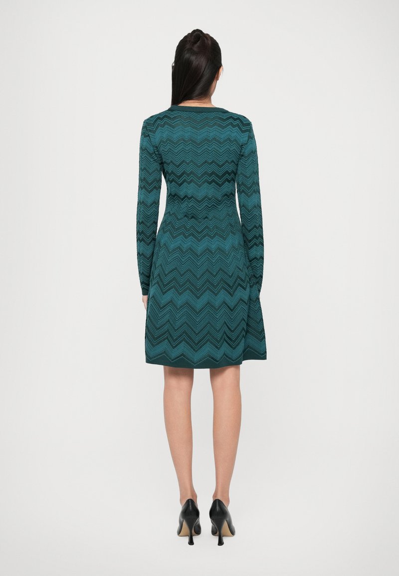 Robe teal avec un motif en zigzag, manches longues, corsage ajusté, jupe évasée et fermeture éclair au dos. Associée à des chaussures à talons hauts noires.
