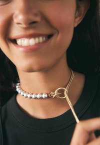 Perlový náhrdelník s chokerom, ozdobený kovovými striebornými korálkami, zlatým kruhovým uzáverom a akcentom v tvare hviezdy. Nosí sa okolo krku v kombinácii s čiernym topom.