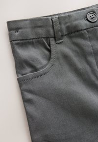 Pantalon en tissu gris à la texture lisse, doté d'une poche avant et d'une fermeture bouton à la taille. Les détails de couture sont visibles.