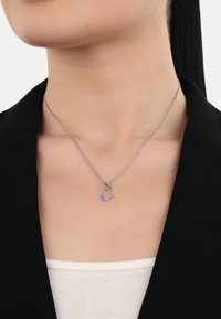 Silberne Anhängerkette mit einem Blumenanhänger auf einer zarten Kette, getragen über einem weißen Oberteil, vor einem schwarzen Jackett.