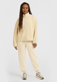 Giacca con zip in pelliccia sintetica e pantaloni coordinati color crema chiaro, caratterizzati da una vestibilità rilassata con polsini elasticizzati e dettagli di cucitura in evidenza.