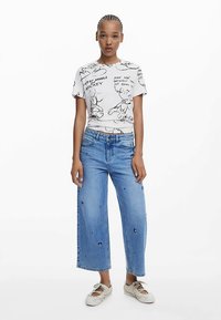 Hvid grafisk t-shirt med sorte illustrationer, kombineret med lyseblå jeans med vide ben og små dekorative patches samt hvide sneakers.