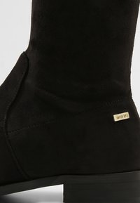 Bottine en daim noire avec un talon bas et une petite plaque métallique rectangulaire avec logo sur le côté.