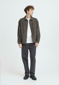 Brun uldblandet jakke med krave, knaplukning, to frontlommer, båret over en hvid skjorte, parret med mørke jeans og sorte sko.