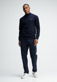 Marinefarbener Rollkragenpullover mit geripptem Saum, kombiniert mit marineblauen Hosen und weißen Sneakers. Weiche Textur, minimales Branding auf der Brust.