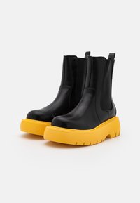 Botas de cuero negras hasta el tobillo con paneles laterales elásticos y suelas de goma gruesa y amarilla brillante sobre un fondo blanco.