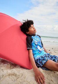 Blå rashguard med sorte accenter, med print af en figur; matchende blå badeshorts med design; rød strandparasol i baggrunden.