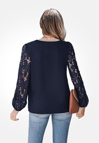Navy blouse with solid back and sheer lace sleeves featuring floral patterns. Paired with light blue jeans and a brown clutch.