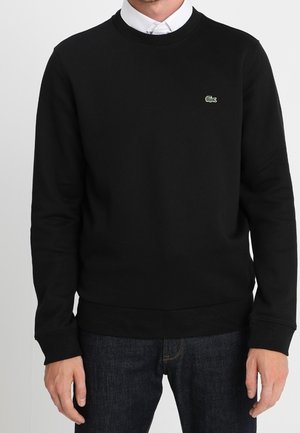 Mężczyzna ubrany w czarną bluzę z okrągłym dekoltem i małym zielonym logo krokodyla na lewej piersi, w połączeniu z ciemnoniebieskimi dżinsami i białą koszulą z kołnierzykiem.