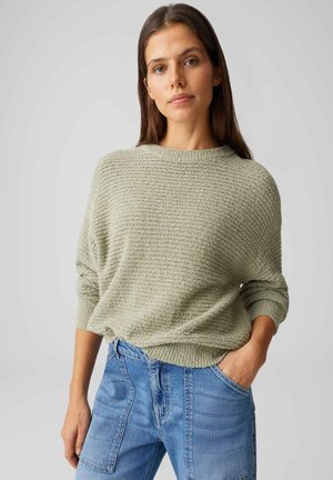 Vrouw met lang bruin haar, gekleed in een lichtgroene, gestructureerde trui en blauwe jeans met hoge taille, staand met één hand in de zak.