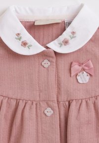 Robe tricotée rose avec un col blanc à broderies florales, fermetures à boutons, accent en nœud en tissu et jupe froncée.