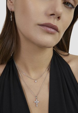 Frau trägt mehrere silberne Halsketten mit einem kleinen runden Anhänger und einem Kreuz mit Edelsteinen sowie einem passenden hängenden Ohrring mit klaren Steinen.