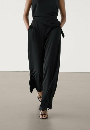 Woman wearing loose black wide-leg pants, black sleeveless top, and black strappy sandals, walking with hands in pockets.