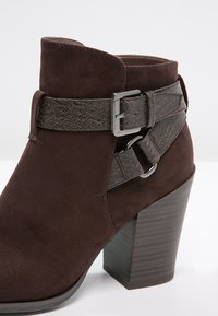 Bota de tobillo de ante marrón con una correa texturizada marrón y detalle de hebilla plateada, que presenta un tacón de madera grueso.