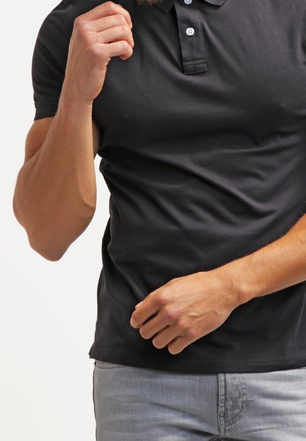 Homme en polo noir et jean gris clair, la main droite levée près de la poitrine et la main gauche près de la taille.