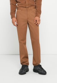 Brown cotton trousers with a straight cut, featuring side pockets and a button closure. Paired with black textured sneakers.