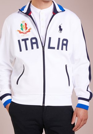 Witte jas met ritssluiting met tekst ITALIA, Italiaanse wapenspreuk en Polo Ralph Lauren-logo, gedragen over een wit overhemd met zwarte broek.