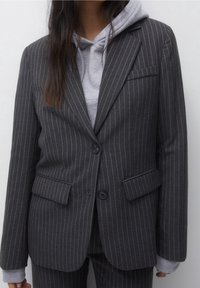 Blazer de rayas gris oscuro con dos bolsillos frontales, dos botones y un solo bolsillo en el pecho, superpuesto a una sudadera con capucha gris.