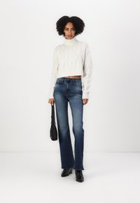 Hvit kabelstrikket kort jakke, blå flared jeans og svarte ankelstøvler. Outfitet er kombinert med en liten svart håndveske med et logo.