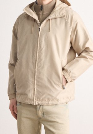 Personne portant une veste à capuche beige zippée par-dessus un pull zippé couleur sable et un jean beige, avec une main dans la poche de la veste.