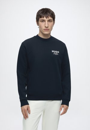 Junger Mann, trägt einen schwarzen HUGO-Sweatshirt und weiße Hose, steht vor einem schlichten weißen Hintergrund und schaut nach vorne.