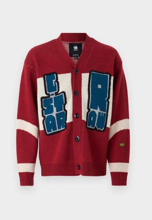 Röd stickad cardigan med v-ringning, med överdimensionerad blå och vit grafisk text. Fem svarta knappar och randiga detaljer på ärmarna.