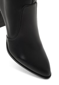 Bottine en cuir noir avec un bout pointu, texture lisse et un talon carré, présentant des détails de couture sur les côtés.