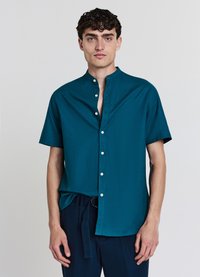 Chemise bleu sarcelle à manches courtes avec col mao, boutons sur le devant et texture lisse. Associée à un pantalon foncé avec une ceinture nouée.